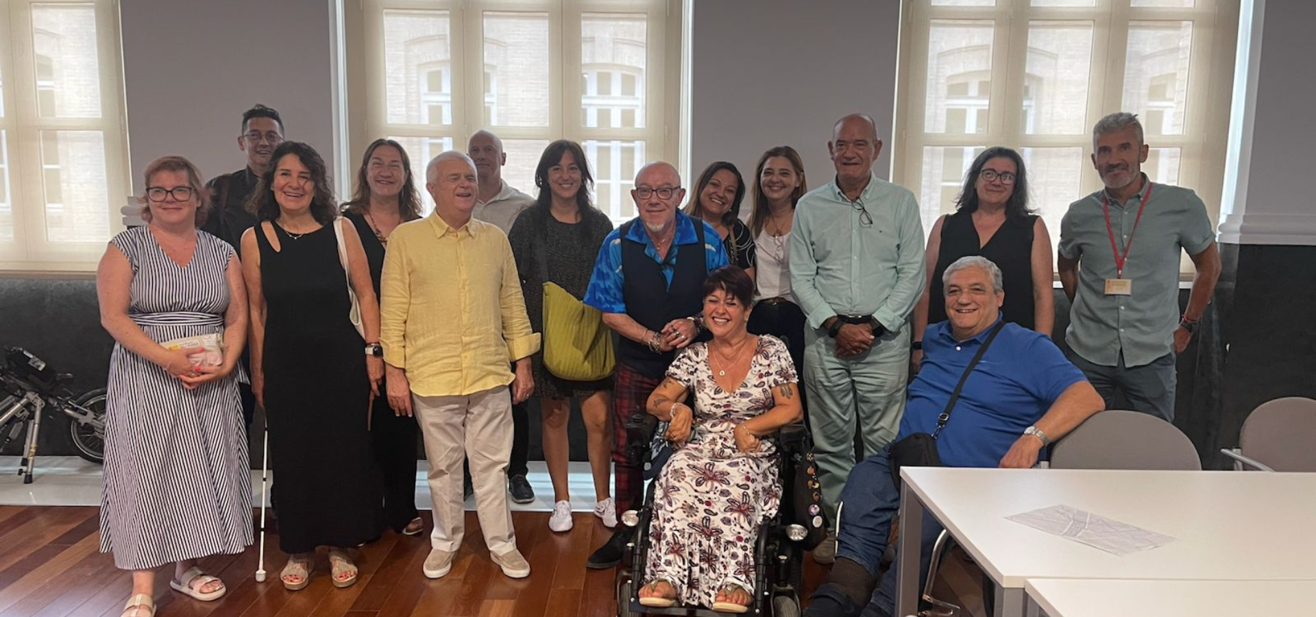 Grupo de personas representantes del ámbito de la discapacidad posando juntas en una sala con suelo de madera y ventanas altas. En el centro, Gemma Mas, en silla de ruedas, sonríe junto a otros miembros del Consejo Municipal de Personas con Discapacidad de València y del CERMI CV, tras la aprobación de su nombramiento como Defensora de las Personas con Discapacidad. La imagen refleja un ambiente cordial e inclusivoo.