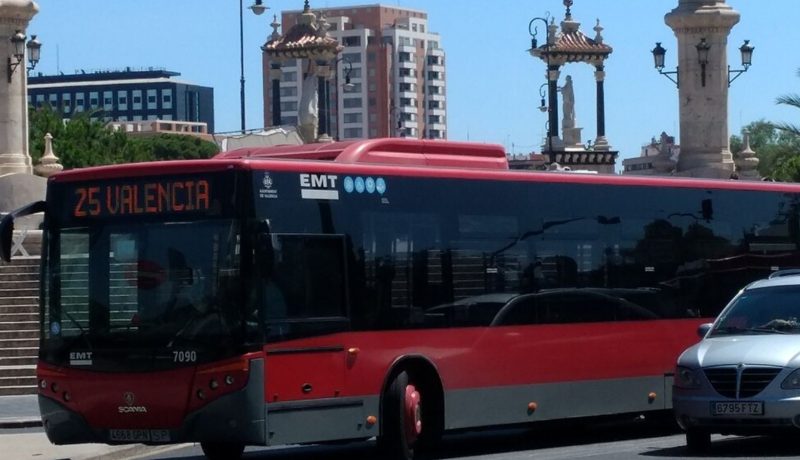 "Autobús urbano de la EMT de Valencia, línea 25 con destino 'Valencia', de color rojo y gris, circulando en la ciudad. A su alrededor se ven coches estacionados, farolas y monumentos en el fondo, bajo un cielo despejado."