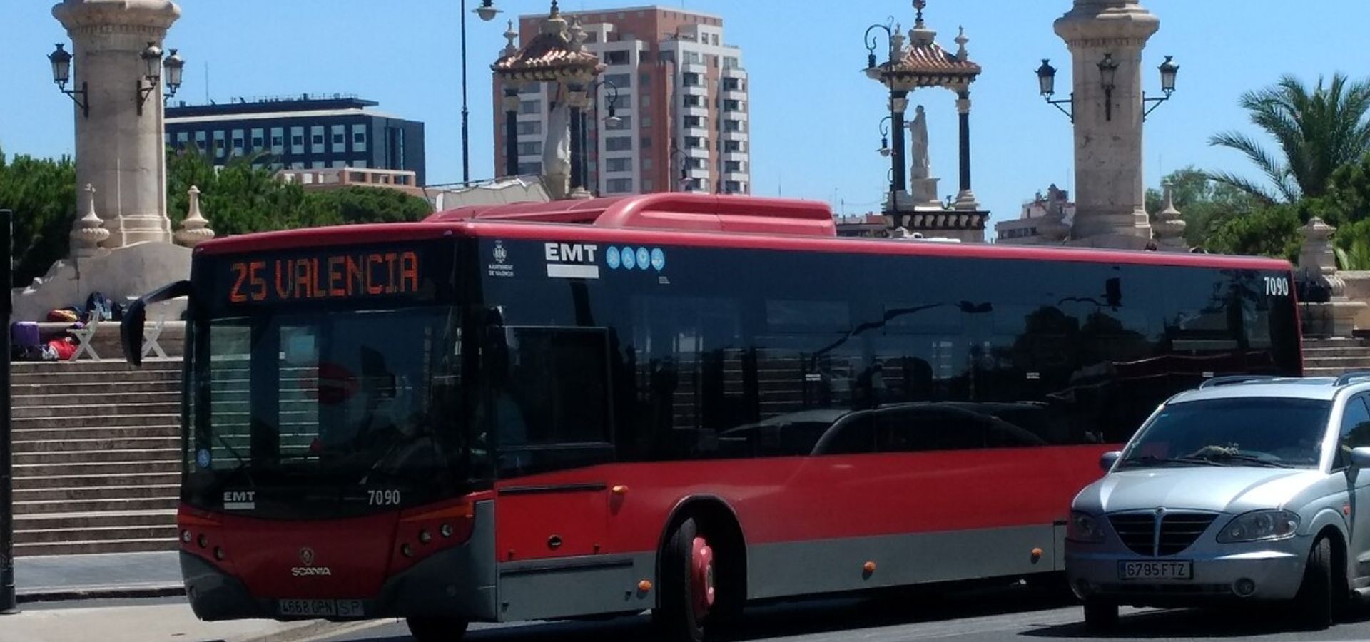 "Autobús urbano de la EMT de Valencia, línea 25 con destino 'Valencia', de color rojo y gris, circulando en la ciudad. A su alrededor se ven coches estacionados, farolas y monumentos en el fondo, bajo un cielo despejado."